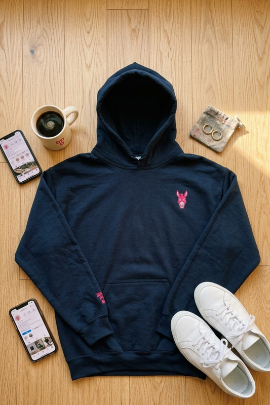 Flat lay of STBN Stubborn by Nature women's navy hoodie on warm wood surface with pink embroidered donkey chest logo visible, styled with coffee mug, white sneakers, two phones and a linen pouch with gold rings.