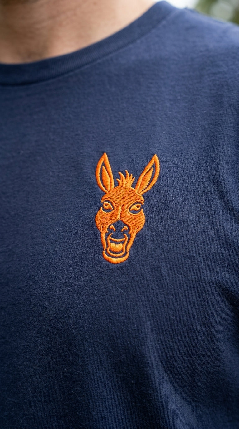 A macro, high-resolution close-up shot of a man's shoulder and chest area. He is wearing a dark navy organic cotton t-shirt. The focus is on the intricate detail of a vibrant orange embroidered donkey logo on the left chest. You can see the individual threads of the embroidery and the soft grain of the high-quality cotton fabric. Natural outdoor daylight. This should look like a premium product review photo.