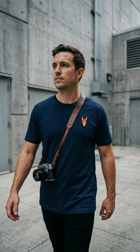 A candid, low-angle street photography shot of a man walking through a clean, industrial urban area. He is wearing a navy blue classic-fit t-shirt. On the left chest is a small, high-density orange embroidered logo of a donkey. The man has a camera strap over his shoulder. The background is a textured grey concrete wall with a slight blur (bokeh). The photo looks like a lifestyle shot from a streetwear blog, realistic lighting, slightly moody city vibes